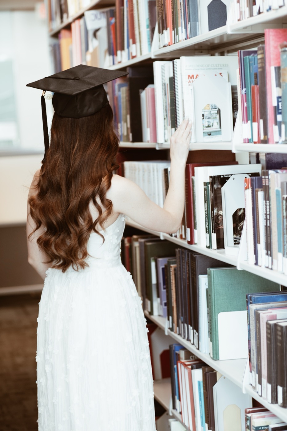 Sophia in cap and gown, browsing the shelves of a library